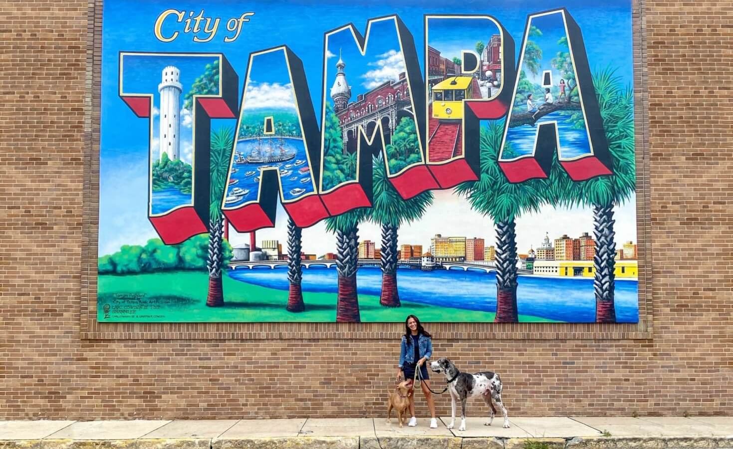 A woman and her two dogs stand in front of a City of Tampa mural