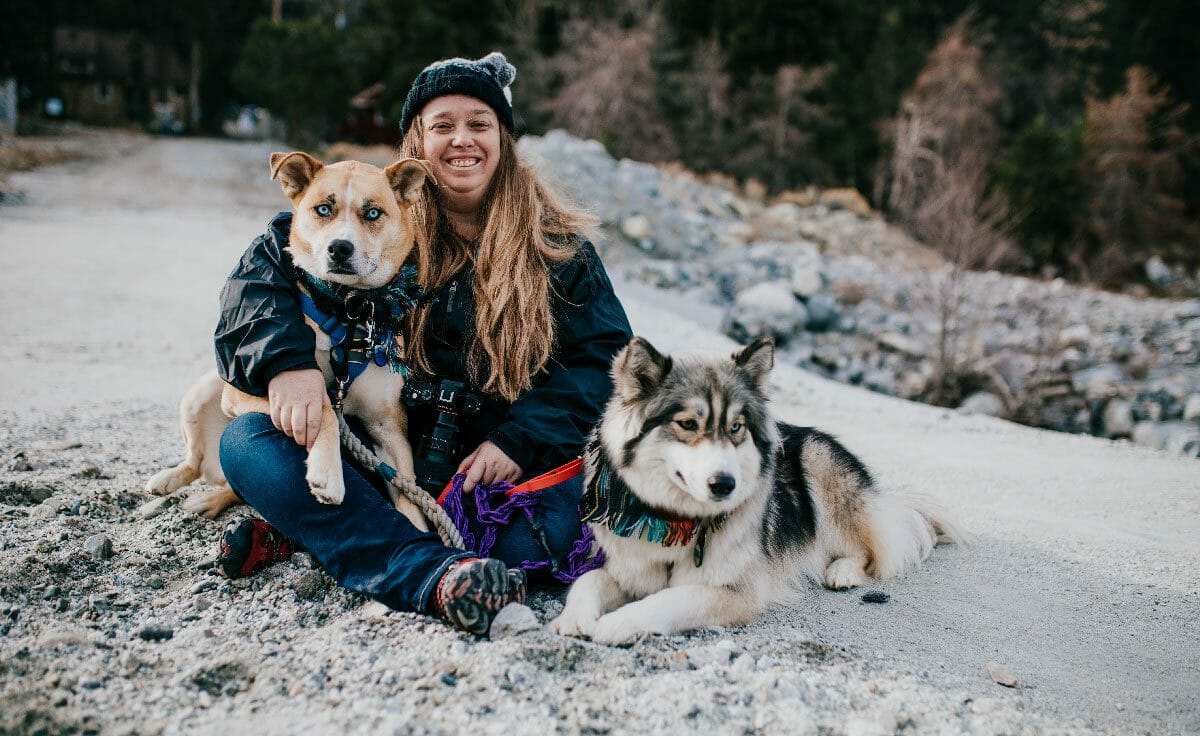 A woman and her two dogs sit together on the ground on a hike in California