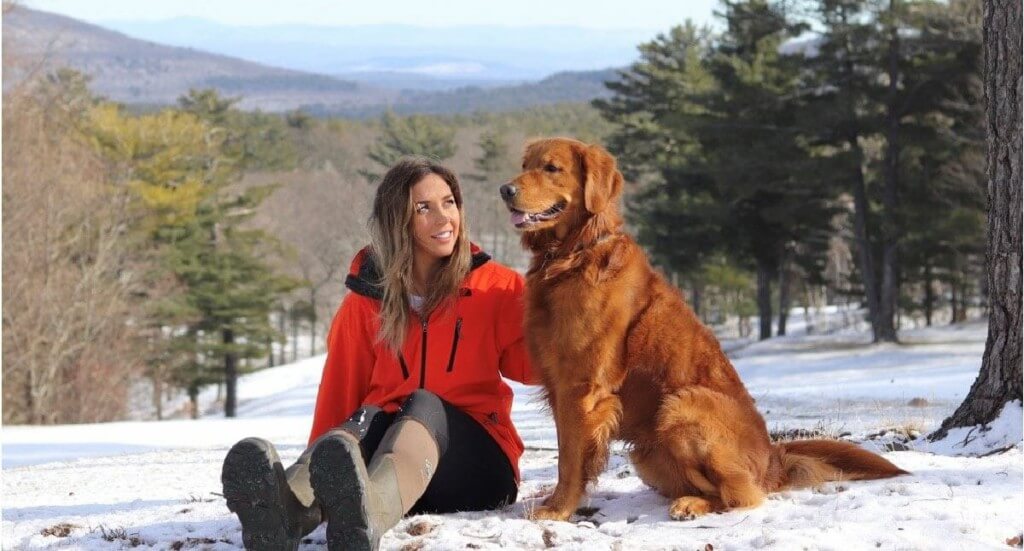 A female in a red coat sits next to her golden retriever dog in the snow with a view of mountains