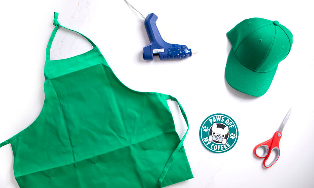 A flat lay of a green apron, hat, hot glue gun, scissors, and a coffee logo