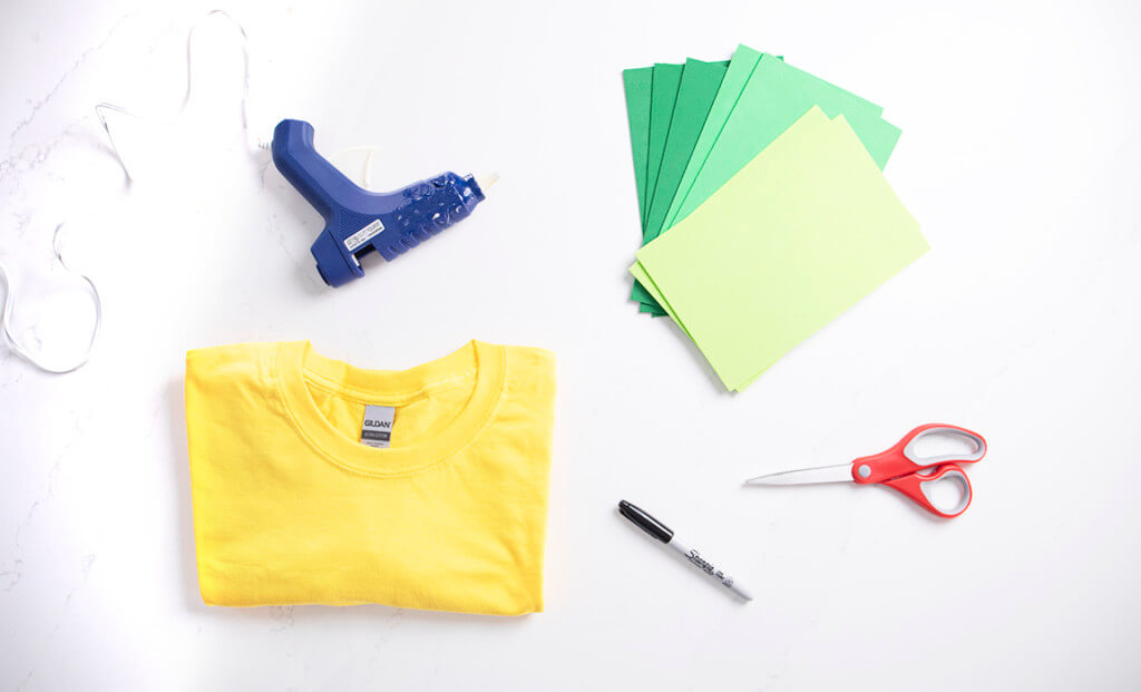 A flat lay of a yellow t-shirt, green foam, a glue gun, scissors, and a marker for a DIY pineapple Halloween costume