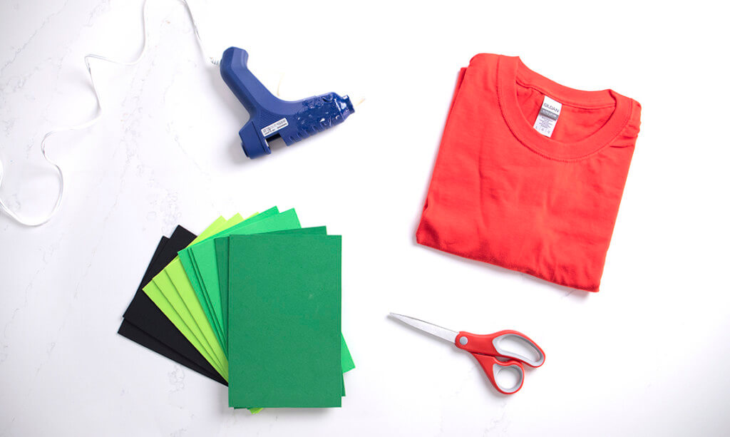 A flat lay of a red t-shirt, black and green foam, a hot glue gun, and scissors for a DIY strawberry Halloween costume