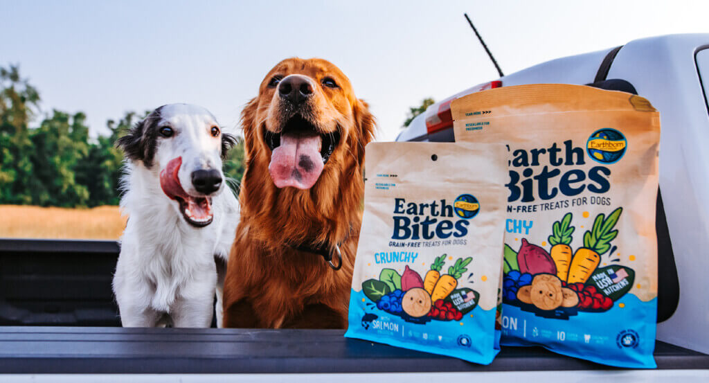 Two dogs sit in back of truck with two bags of EarthBites Crunchy with Salmon Protein