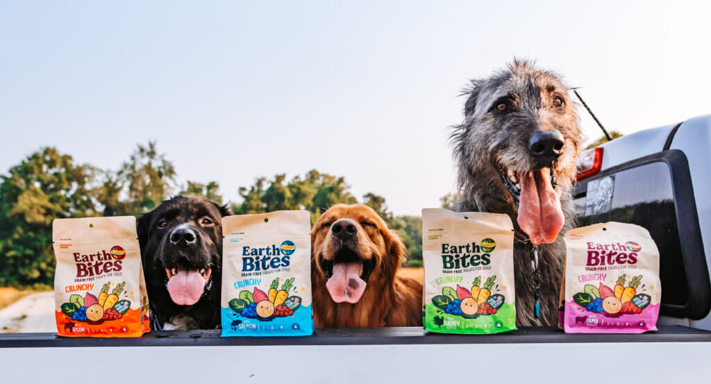 Three dogs sit in back of truck with 4 bags of EarthBites Crunchy lined up on the side of truck.