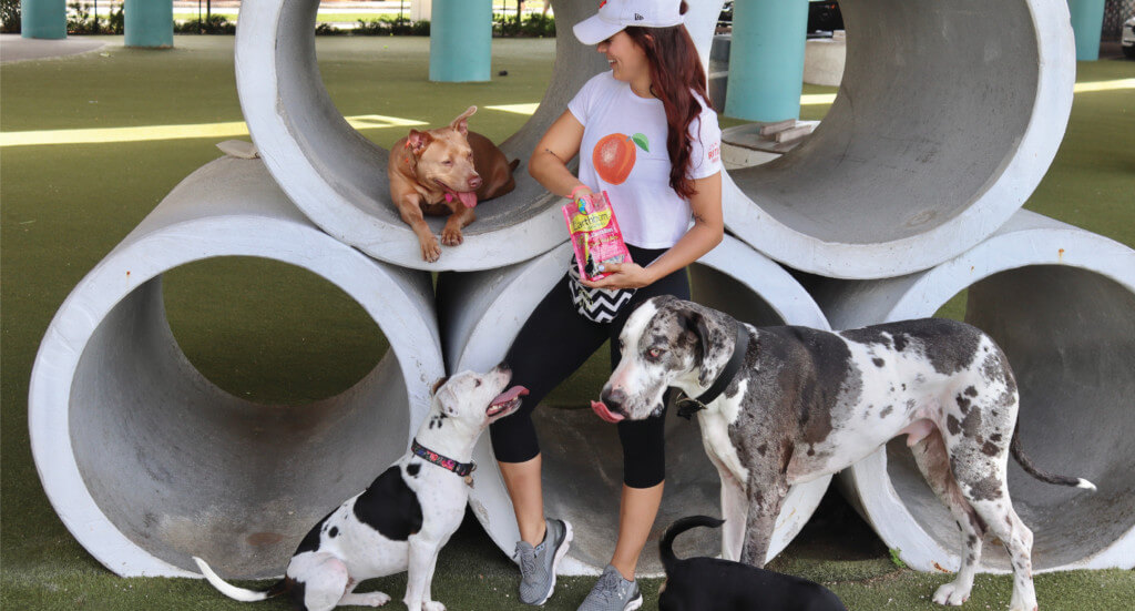 A woman stands in a park with a bag of treats surrounded by dogs