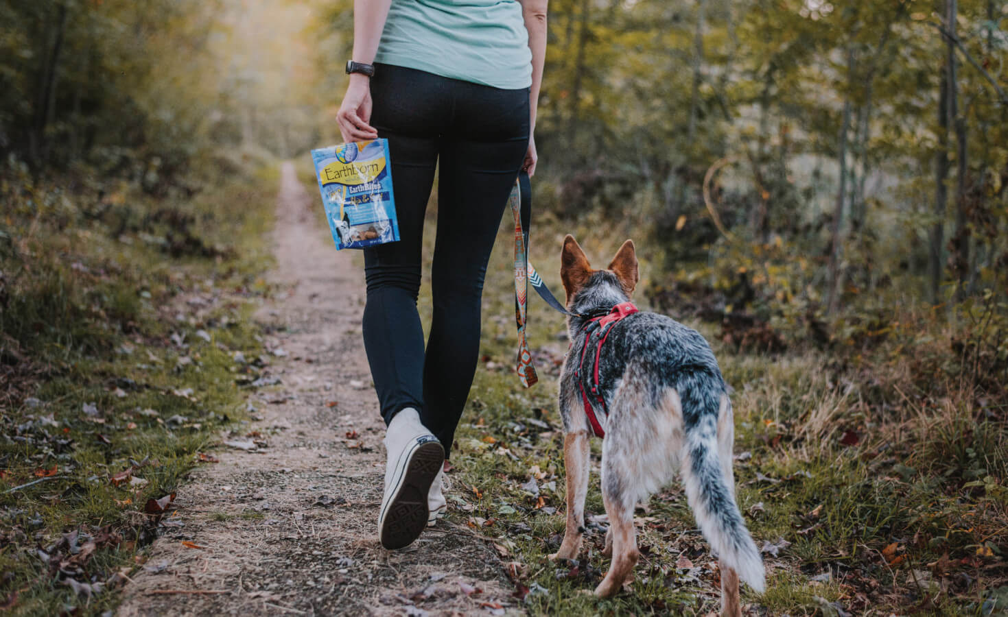 A woman walks her dog on a wooded trail with a bag of EarthBites dog treats in her hand