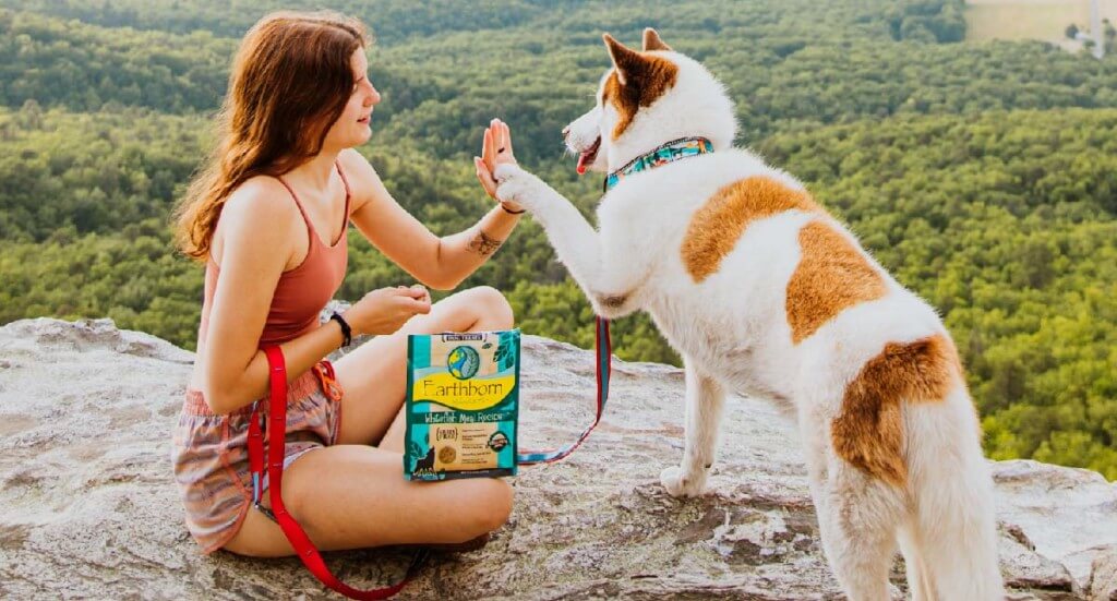 A woman sits with her dog at a lookout spot on a mountain. The dog is giving her a high-five and she has Earthborn Holistic dog treats in her lap