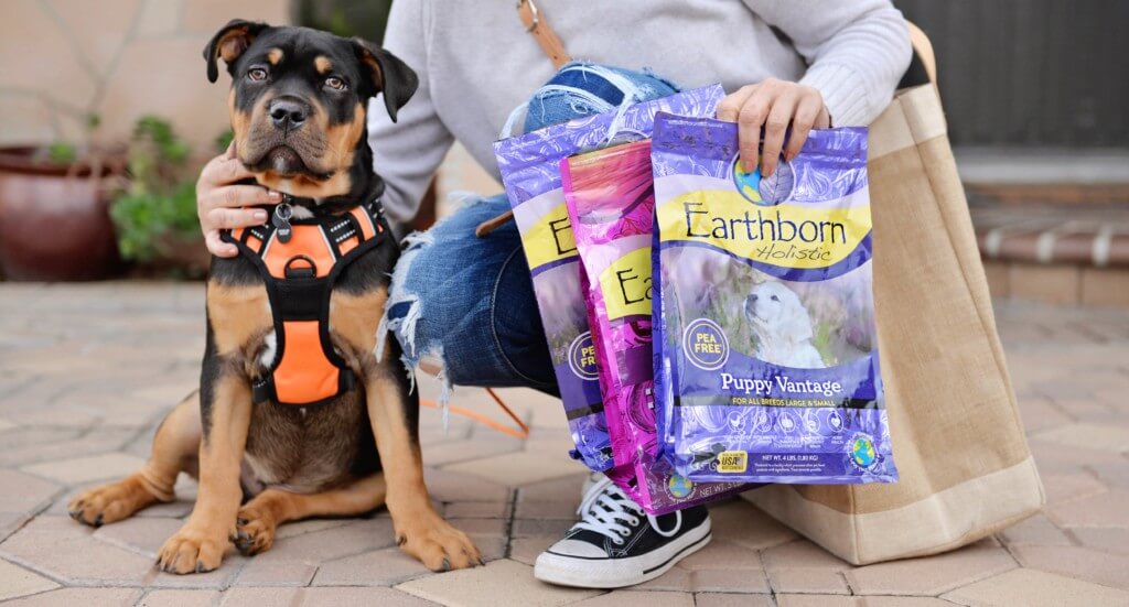 A puppy sits next to his owner who is holding empty Earthborn Holistic dog food bags