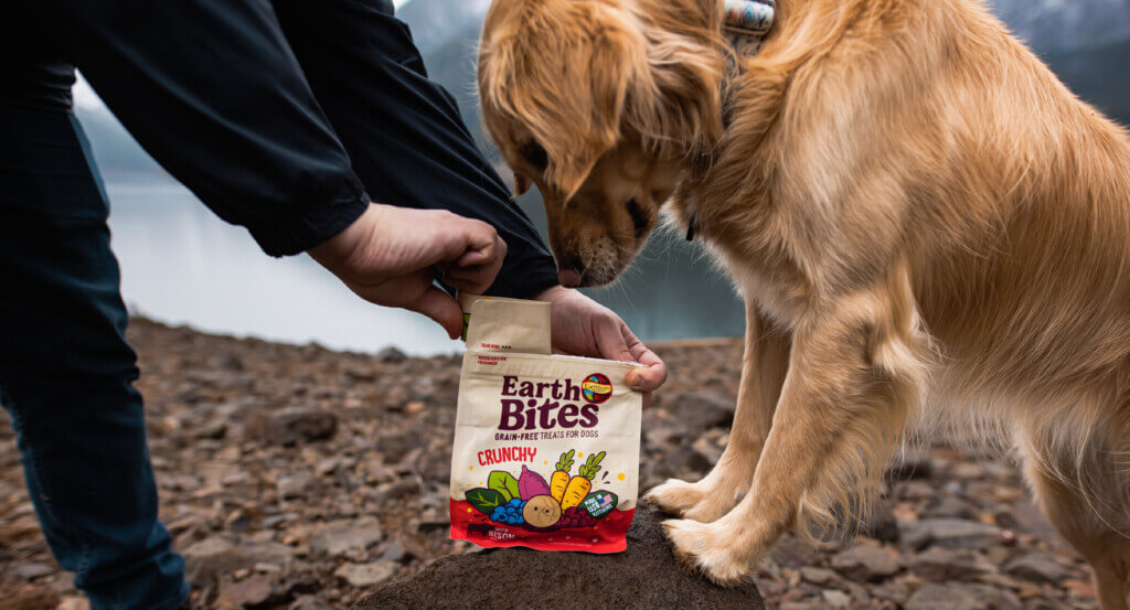 Person holds bag of EarthBites Crunchy treats open as dog looks inside bag