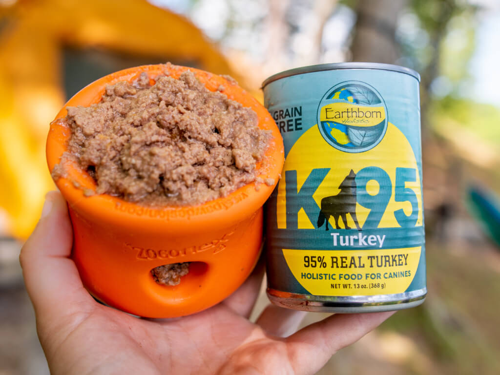 A hand holds both a can of K95 Turkey dog food and an enrichment dog toy filled with the food