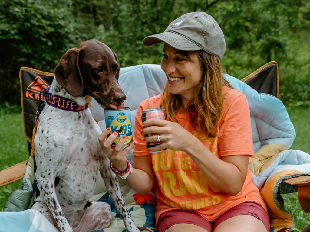 A woman and her dog are sitting in a camping chair. She's holding a canned beverage and holding a can of K95 dog food up for her dog to lick out of