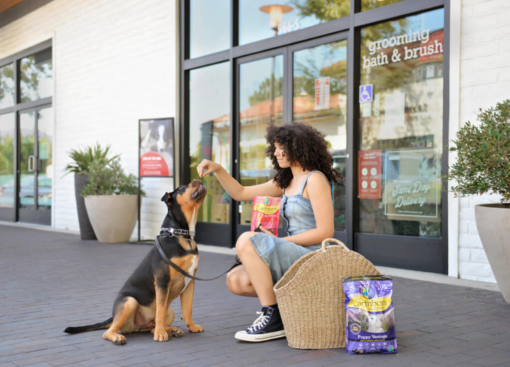 A girl crouches on the ground with a dog treat bag while a puppy reaches up to get a treat