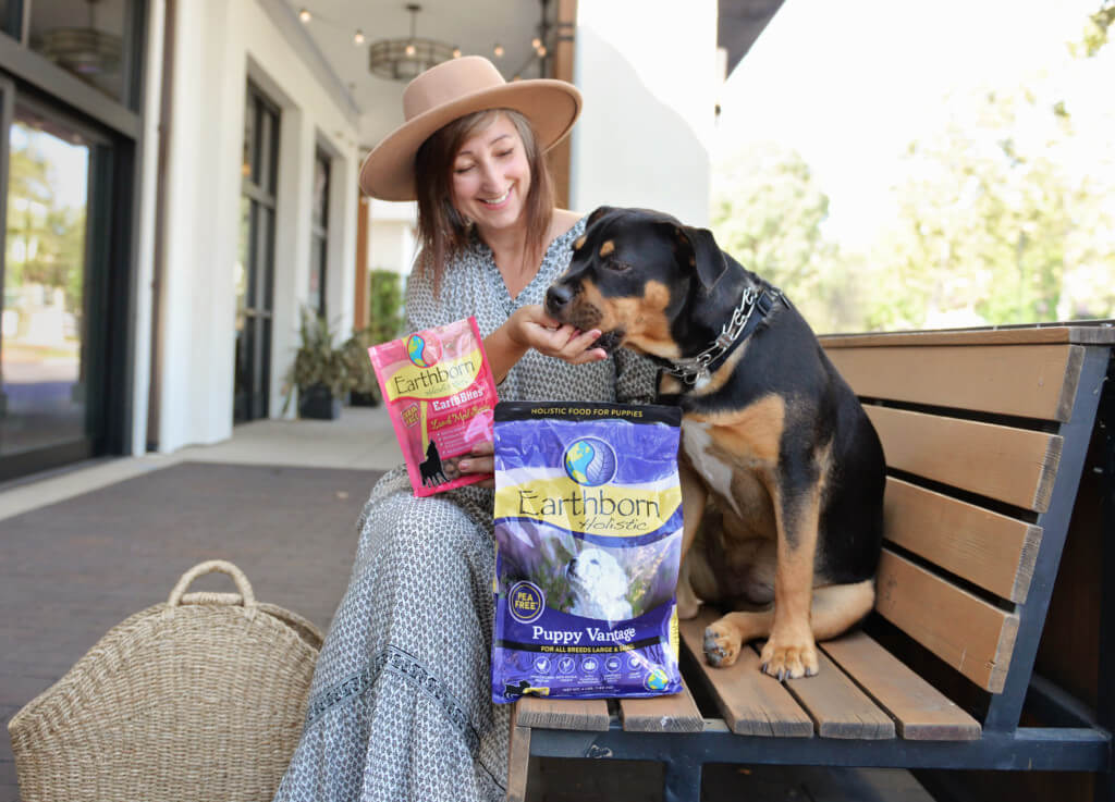 A woman and her puppy sit on an outdoor bench. She's holding a bag of treats and a bag of puppy food is sitting on the bench with them