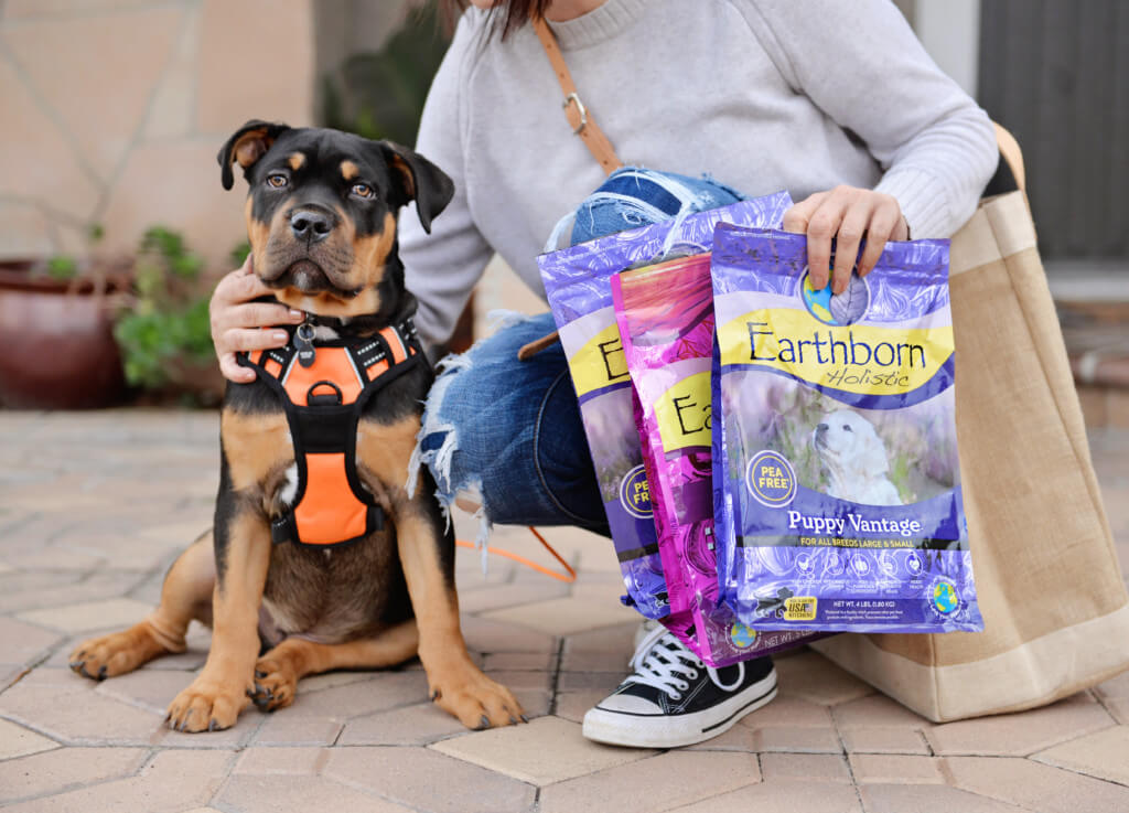A woman crouches on the ground with her puppy while holding empty recyclable dog food bags