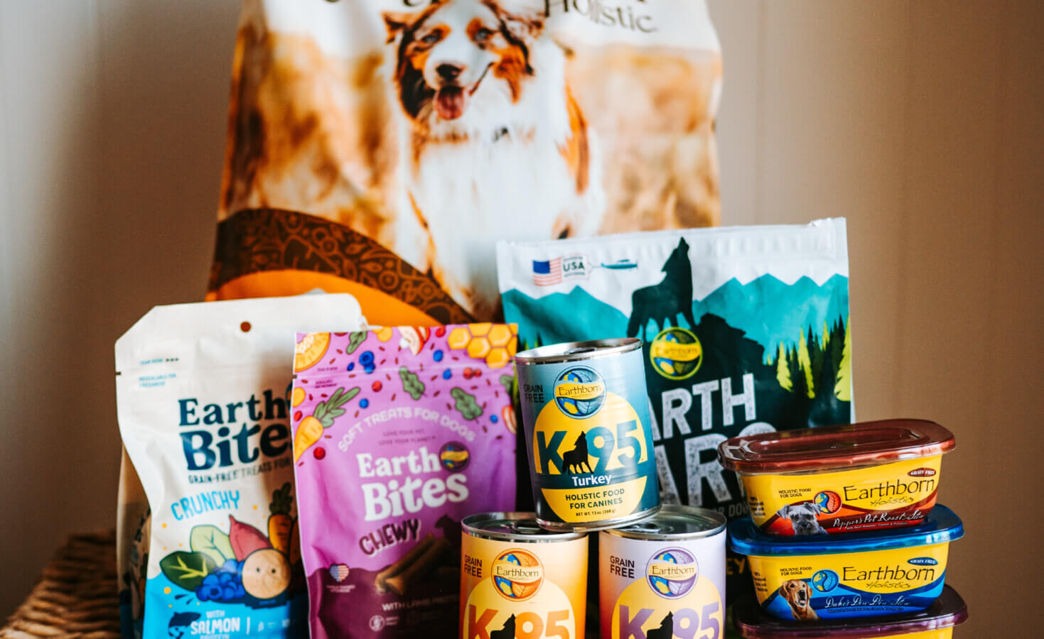 a variety of Earthborn Holistic dry and wet dog food along with treats sits on a basket