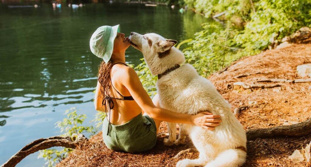 Dog licks woman's face as they sit next to each other on the ground.