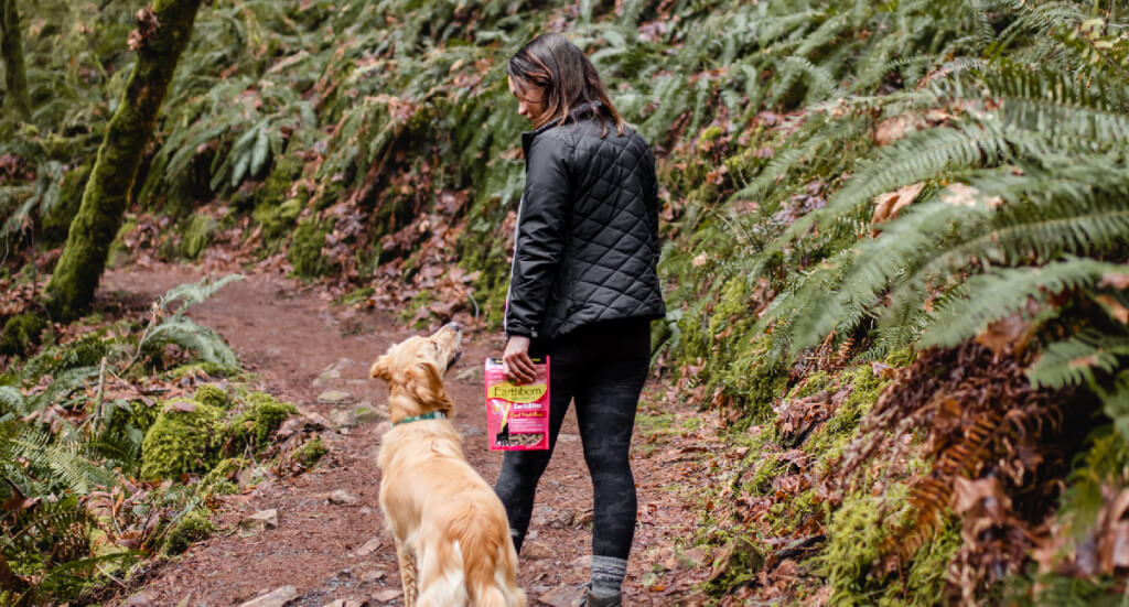 A woman walks with her dog on a forest trail while holding a bag of EarthBites dog treats