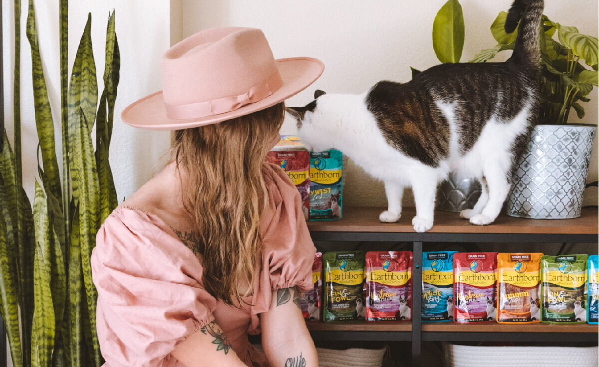 A woman sits in front of a shelf filled with wet cat food pouches while her cat stands on top of the shelf sniffing at a pouch