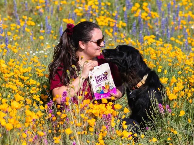 A woman and her dog sit in a field of wildflowers while the woman holds a bag of EarthBites Crunchy Lamb Recipe dog treats