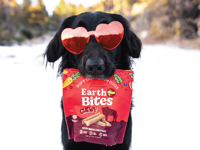 black dog with heart sunglasses holding a red bag of EarthBites Chewy treats
