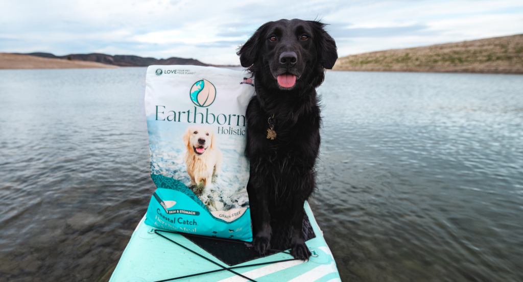 A black dog on a kayak on water with a bag of Earthborn Holistic Sensitive Skin and Stomach Costal Catch.